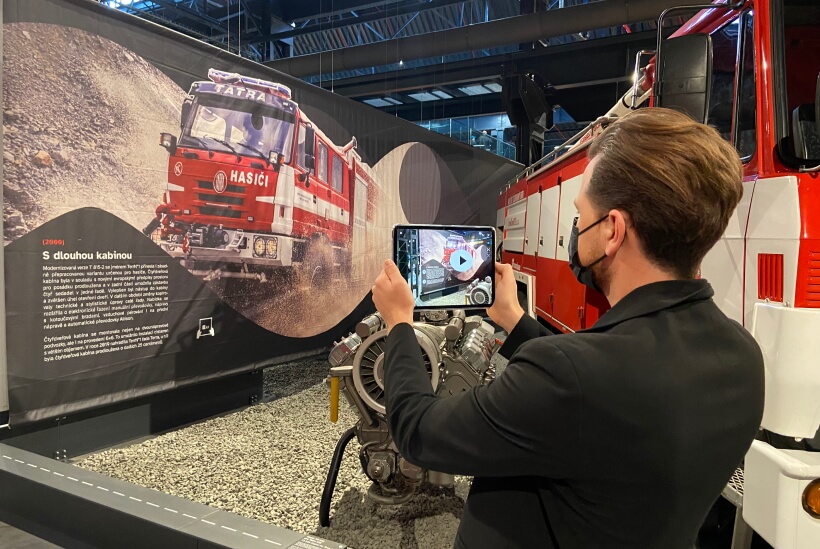 A visitor at an AR museum using a smartphone to interact with a virtual reality display of a Tatra fire truck exhibit